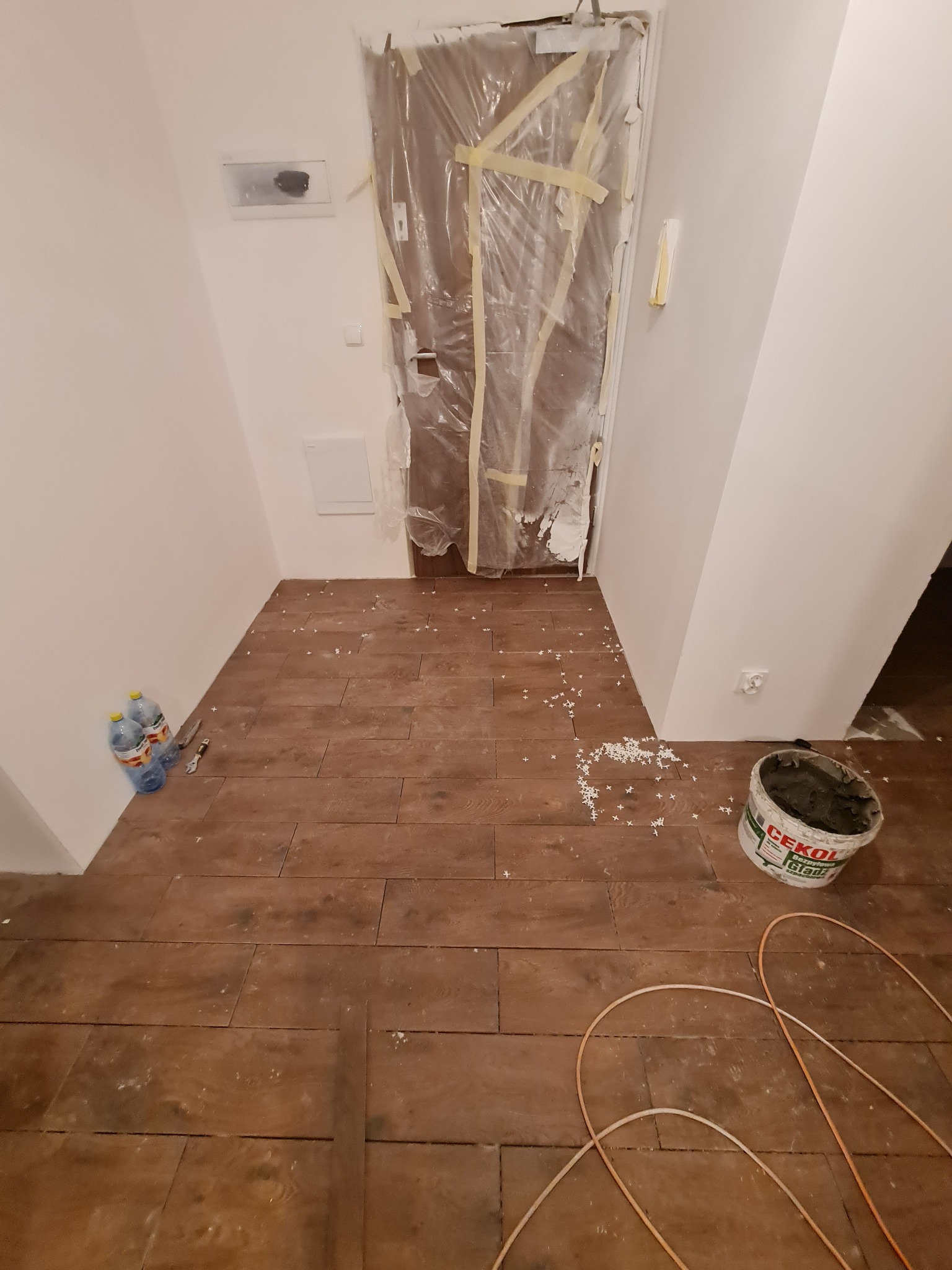 Ułożona podłoga z płytek imitujących drewno w korytarzu, zabezpieczone drzwi folią, wiadro z klejem i krzyżyki dystansowe. Widok z góry na etap prac wykończeniowych.