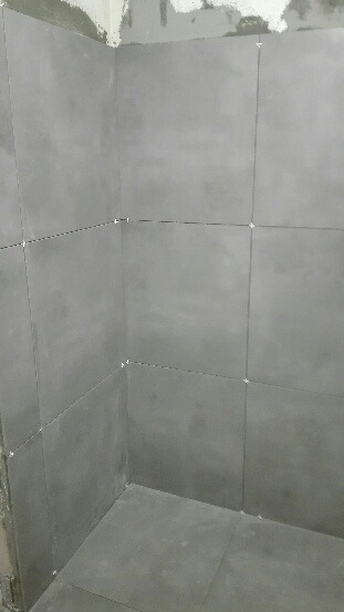 Świeżo ułożone, szare płytki w rogu łazienki, z widocznymi krzyżykami dystansowymi. Surowy beton na górze ściany. Ujęcie z dołu, podkreślające regularność wzoru.