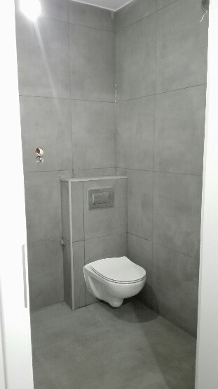 Szara łazienka po remoncie z podwieszaną toaletą i płytkami imitującymi beton. Widoczne przyłącza, minimalistyczny styl.