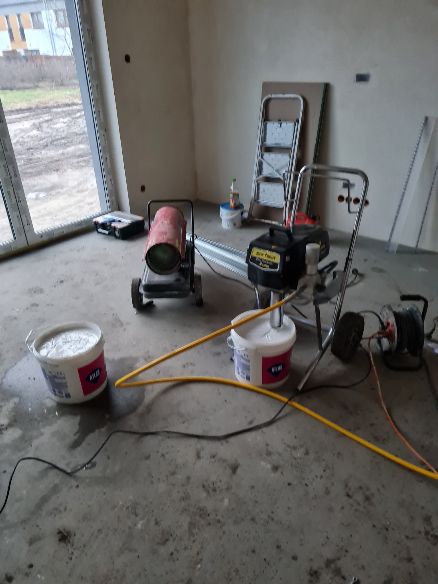 Wnętrze w trakcie malowania maszynowego. Widoczny agregat malarski, wiadro z farbą Atlas, nagrzewnica i drabina. Surowy stan ścian przed ostatecznym wykończeniem.