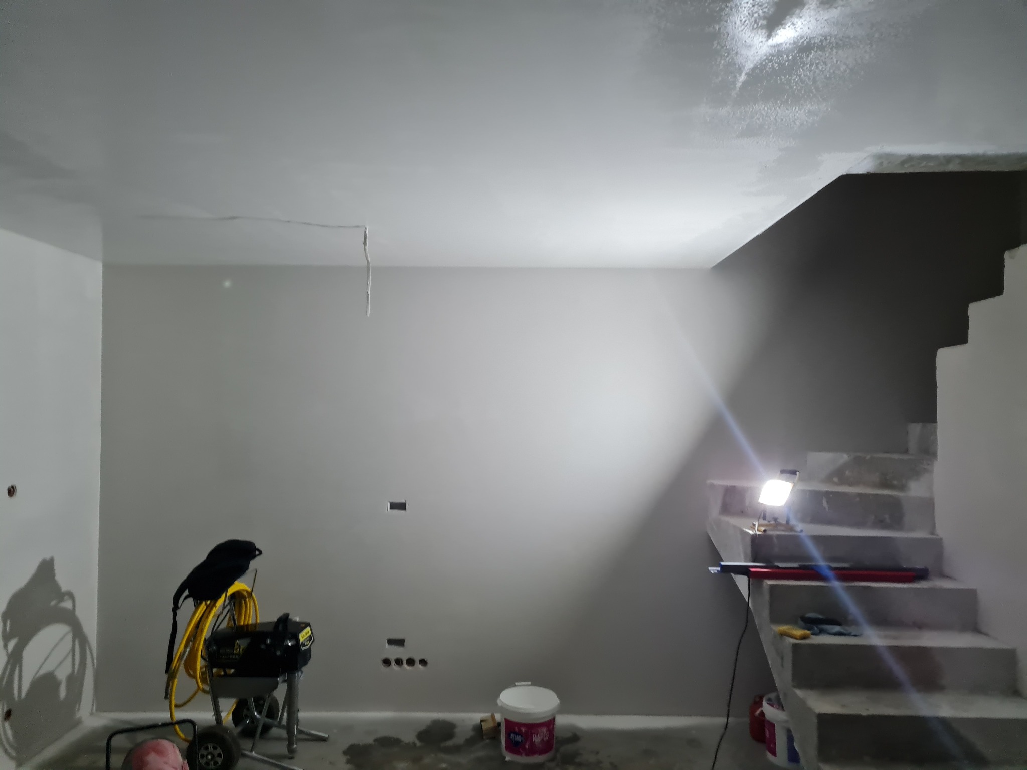 Wnętrze po szpachlowaniu: gładkie, szare ściany, surowe schody z oświetleniem roboczym, agregat malarski z żółtym wężem i wiadro farby. Przygotowanie do malowania.