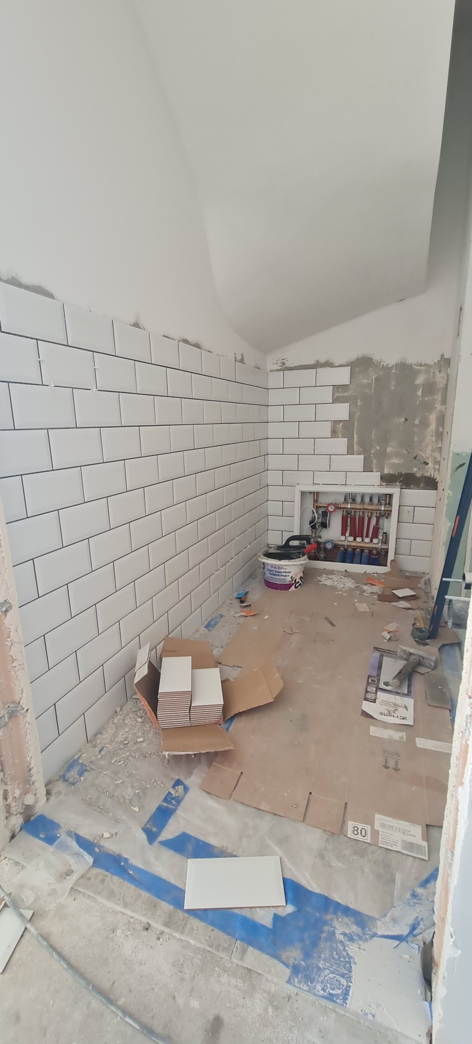 Białe, prostokątne płytki ułożone na ścianie w łazience, częściowo wykończone. Na podłodze kartony i pojedyncze płytki. Widoczna instalacja wodna.