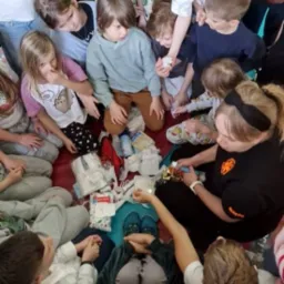 Instruktorka prowadzi kurs pierwszej pomocy dla dzieci, demonstrując opatrywanie ran na fantomie. Dzieci obserwują z bliska, siedząc wokół na podłodze.