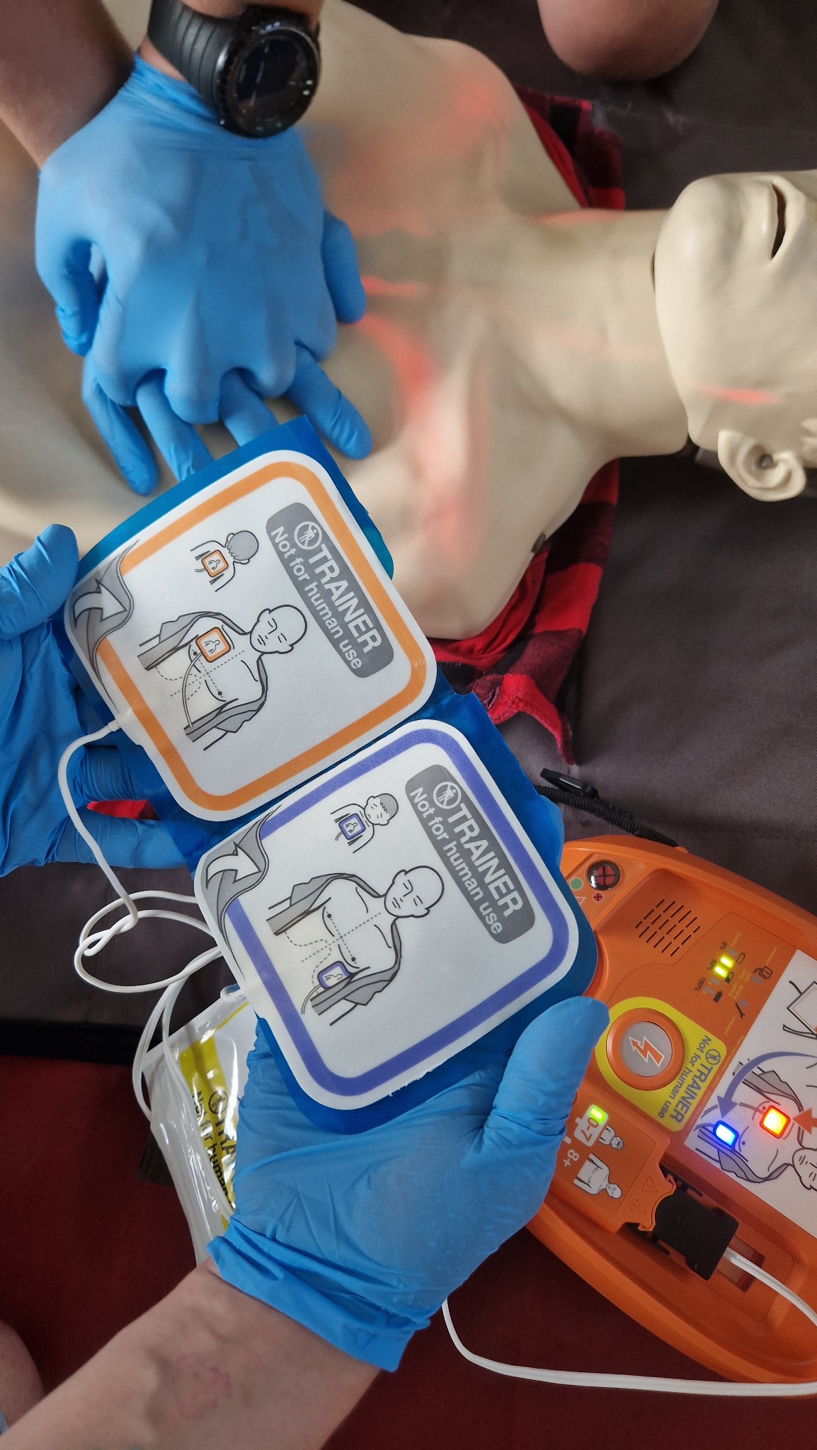 Manekin z podłączonymi elektrodami defibrylatora treningowego, dłonie w niebieskich rękawiczkach w trakcie ćwiczeń z użyciem defibrylatora treningowego.