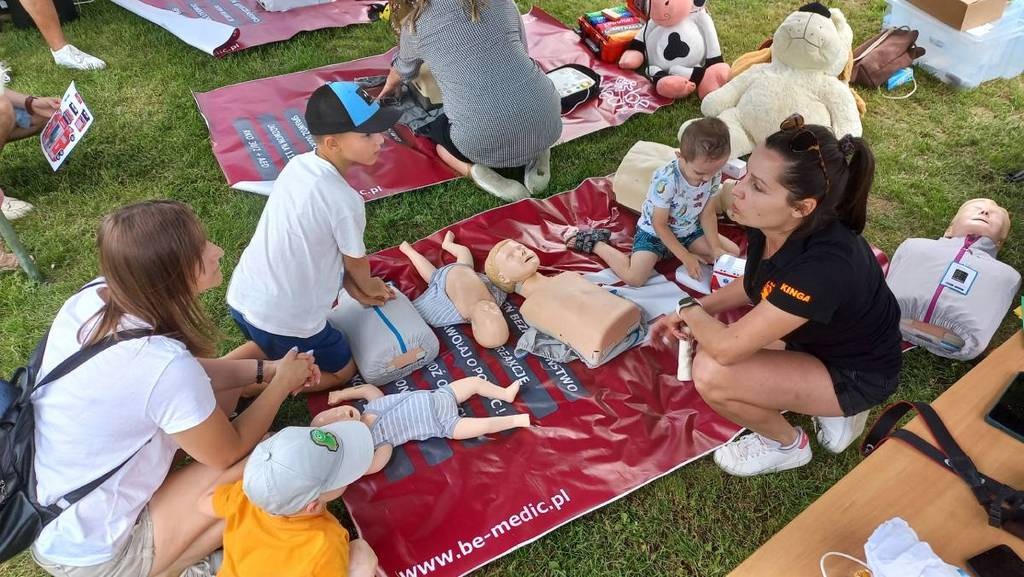 Szkolenie z pierwszej pomocy na trawie: instruktorka demonstruje na manekinie, dzieci obserwują. Czerwony baner z napisem 'be-medic.pl' w tle.