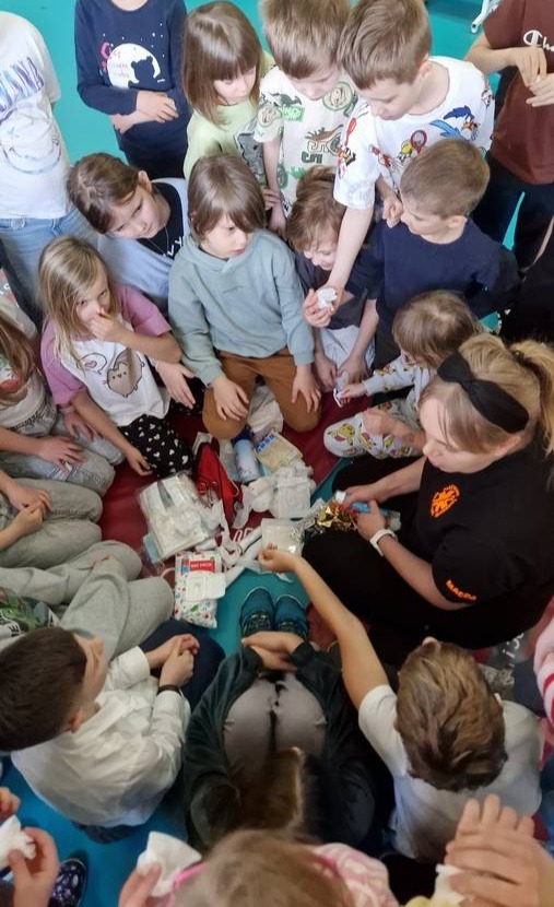 Instruktorka prowadzi kurs pierwszej pomocy dla dzieci, demonstrując opatrywanie ran na fantomie. Dzieci obserwują z bliska, siedząc wokół na podłodze.