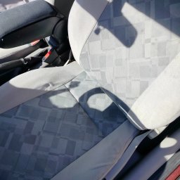 V.P.M.M. Profiti - Szare, materiałowe siedzenie samochodowe z wzorem w kratę, widoczny podłokietnik i pas bezpieczeństwa, oświetlone naturalnym światłem. Czyste i zadbane wnętrze auta.