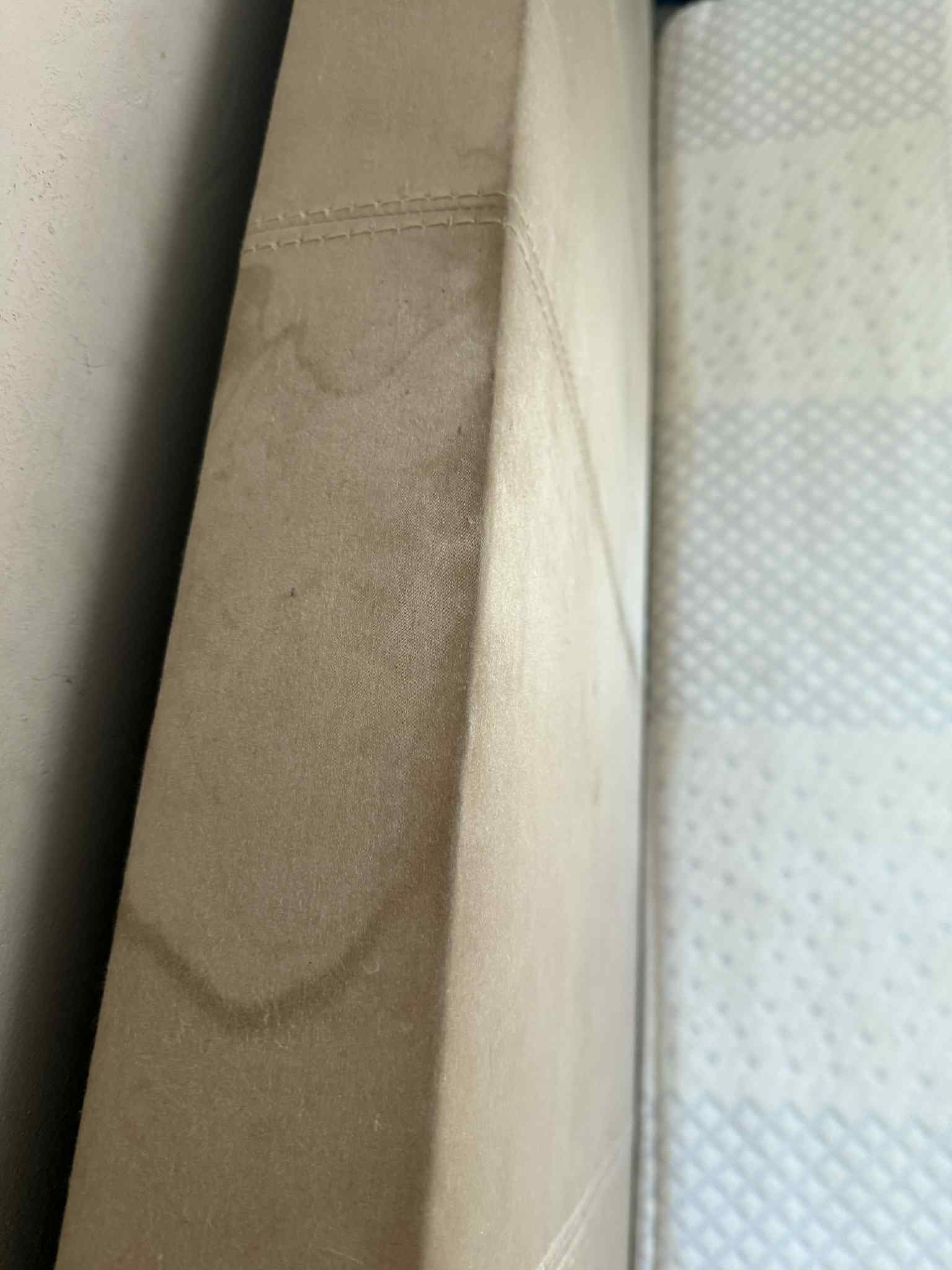 Beżowy narożnik z plamami na tapicerce, widoczne szwy. Obok materac w jasnym odcieniu z delikatnym wzorem. Perspektywa z bliska, ukazująca detale zabrudzeń.