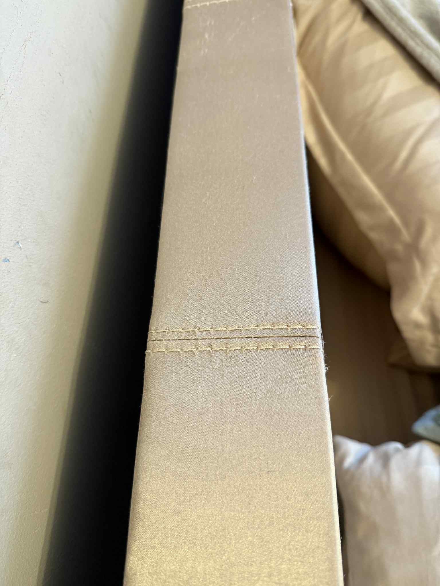 Beżowa tapicerka kanapy z bliska, widoczne szwy i fragment poduszki w tle. Delikatna faktura materiału, neutralna kolorystyka.