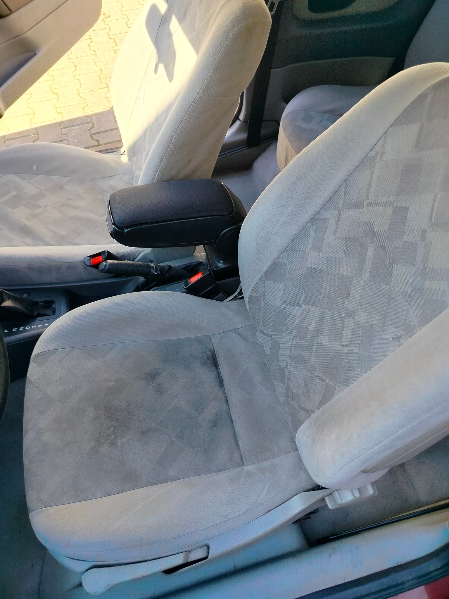 Szare, tekstylne siedzenie samochodowe z widocznymi plamami. Fotel z podłokietnikiem, zapięty pas bezpieczeństwa. Widoczna dźwignia zmiany biegów.