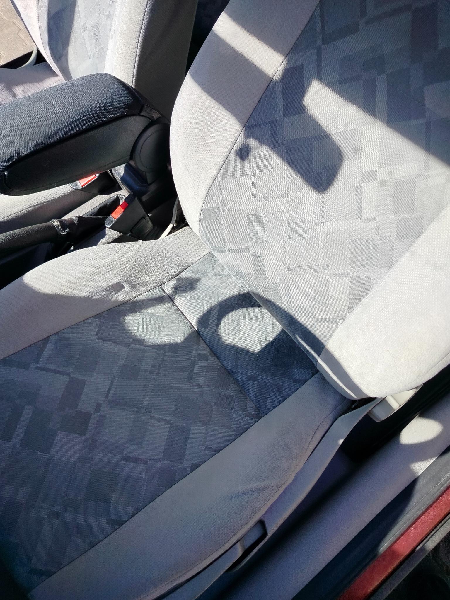 Szare, materiałowe siedzenie samochodowe z wzorem w kratę, widoczny podłokietnik i pas bezpieczeństwa, oświetlone naturalnym światłem. Czyste i zadbane wnętrze auta.