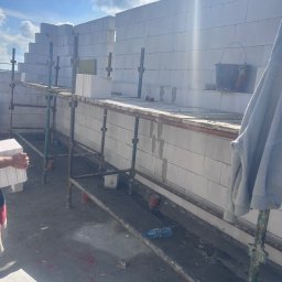 Bob - Wznoszenie ściany z białych bloczków na rusztowaniu. Widoczny fragment ręki z bloczkiem, wiadro i butelka. Słoneczny dzień, budowa w toku.