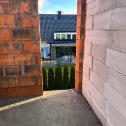 Bob - Wznoszenie ścian domu z pustaków ceramicznych i betonu komórkowego. Widoczny fragment ogrodu z tujami i budynek w tle. Wąskie ujęcie, prezentujące detale konstrukcji.