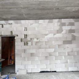 Bob - Ściana z białych bloczków, częściowo wymurowana, widoczne spoiny. W tle fragment ściany z cegły dziurawki i surowy betonowy strop. Na podłodze materiały budowlane.