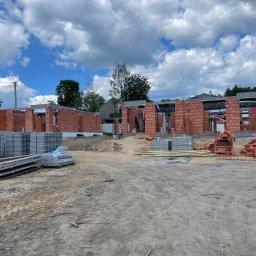 Bob - Wznoszenie murów z czerwonej cegły na budowie domu jednorodzinnego. Widoczne bloczki betonowe i elementy konstrukcyjne na tle błękitnego nieba z chmurami.