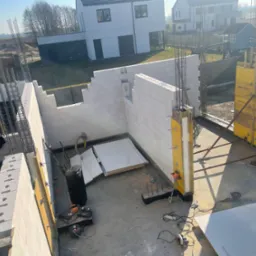 Budowa domu z betonu komórkowego: wzniesione ściany, widoczne zbrojenie i szalunki. Na podłodze narzędzia budowlane, w tle domy jednorodzinne.