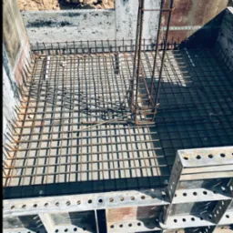 Zbrojenie fundamentu w szalunku stalowym, widok z góry. Stalowe pręty tworzą siatkę, a pionowe elementy zbrojenia wystają ponad nią. Część szalunku widoczna w dolnej części kadru.