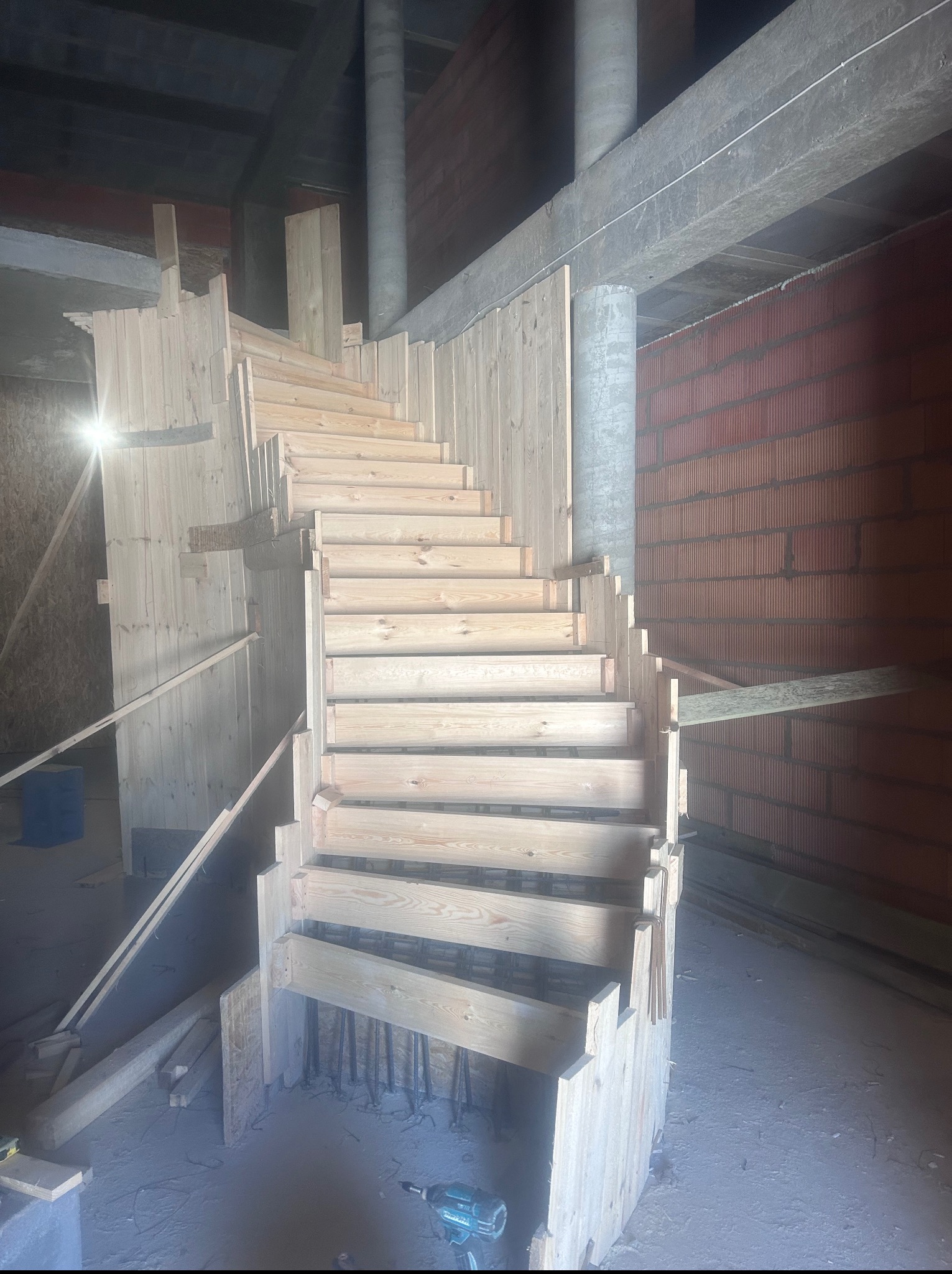 Drewniany szalunek spiralnych schodów na budowie, widoczne zbrojenie i ściany z czerwonej cegły. W tle betonowe słupy, na podłodze wiertarka.