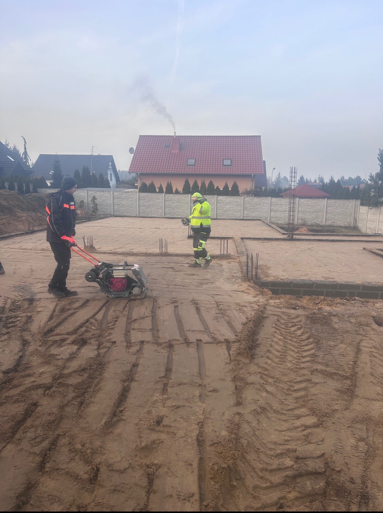 Prace ziemne przy fundamentach: ubijanie gruntu zagęszczarką. Widoczni dwaj pracownicy w odzieży roboczej, przygotowany teren pod budowę domu jednorodzinnego.