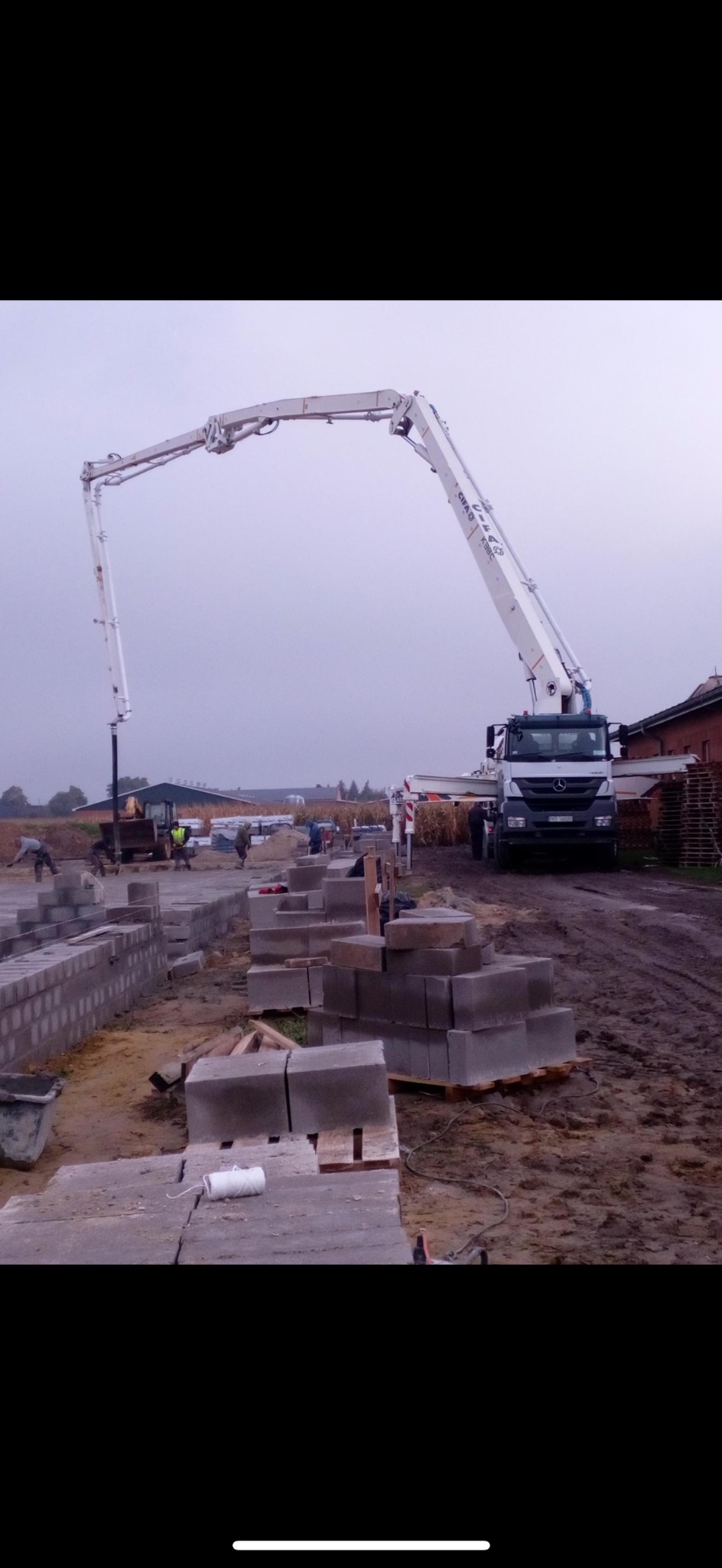 Betonomieszarka pompuje beton na budowę fundamentów z bloczków betonowych. Widoczni pracownicy i sprzęt budowlany w pochmurny dzień.