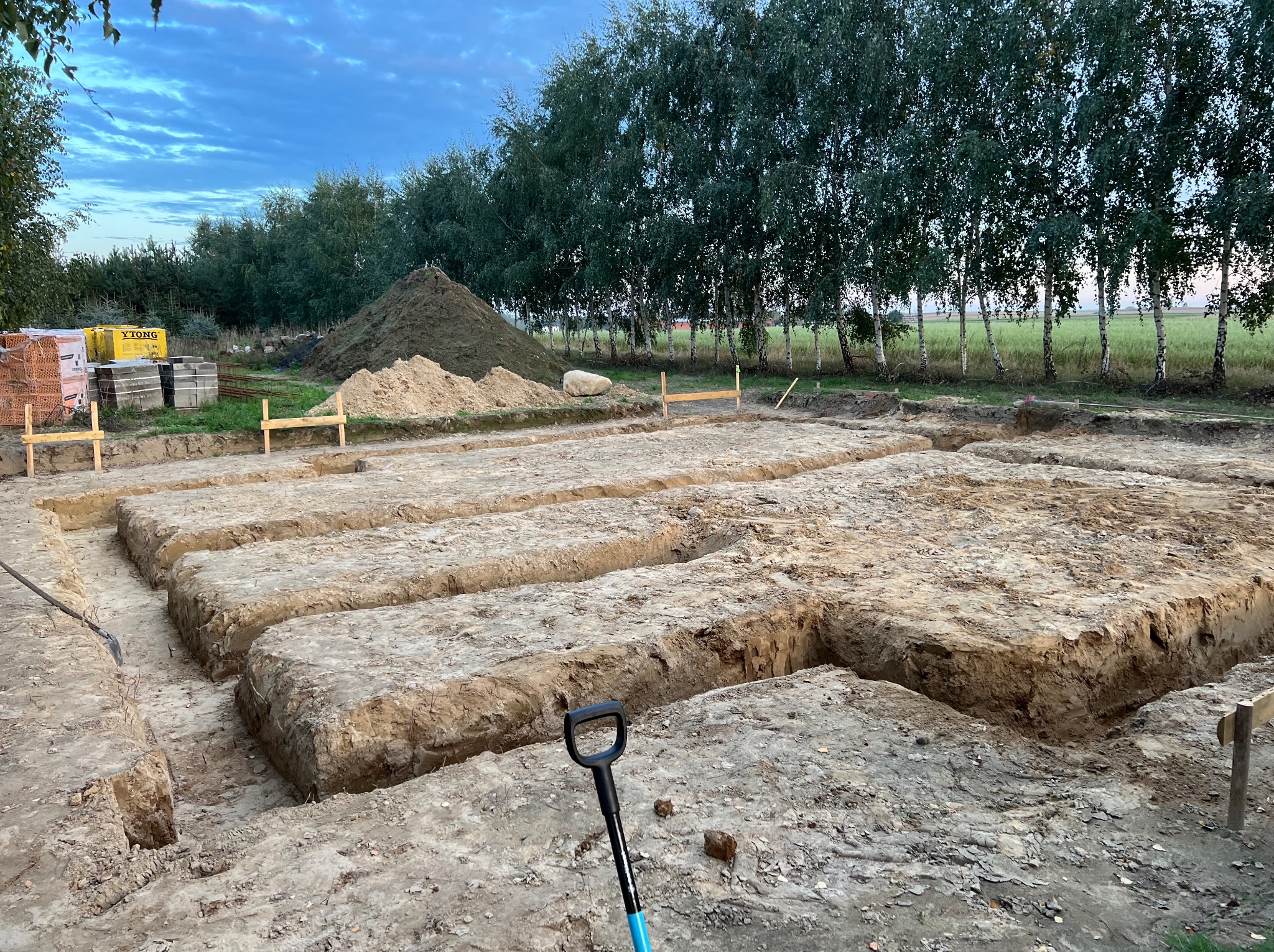 Wykopane rowy pod fundamenty domu jednorodzinnego. Widoczny stos cegieł Ytong i hałda ziemi. W tle rząd drzew i błękitne niebo o zmierzchu.