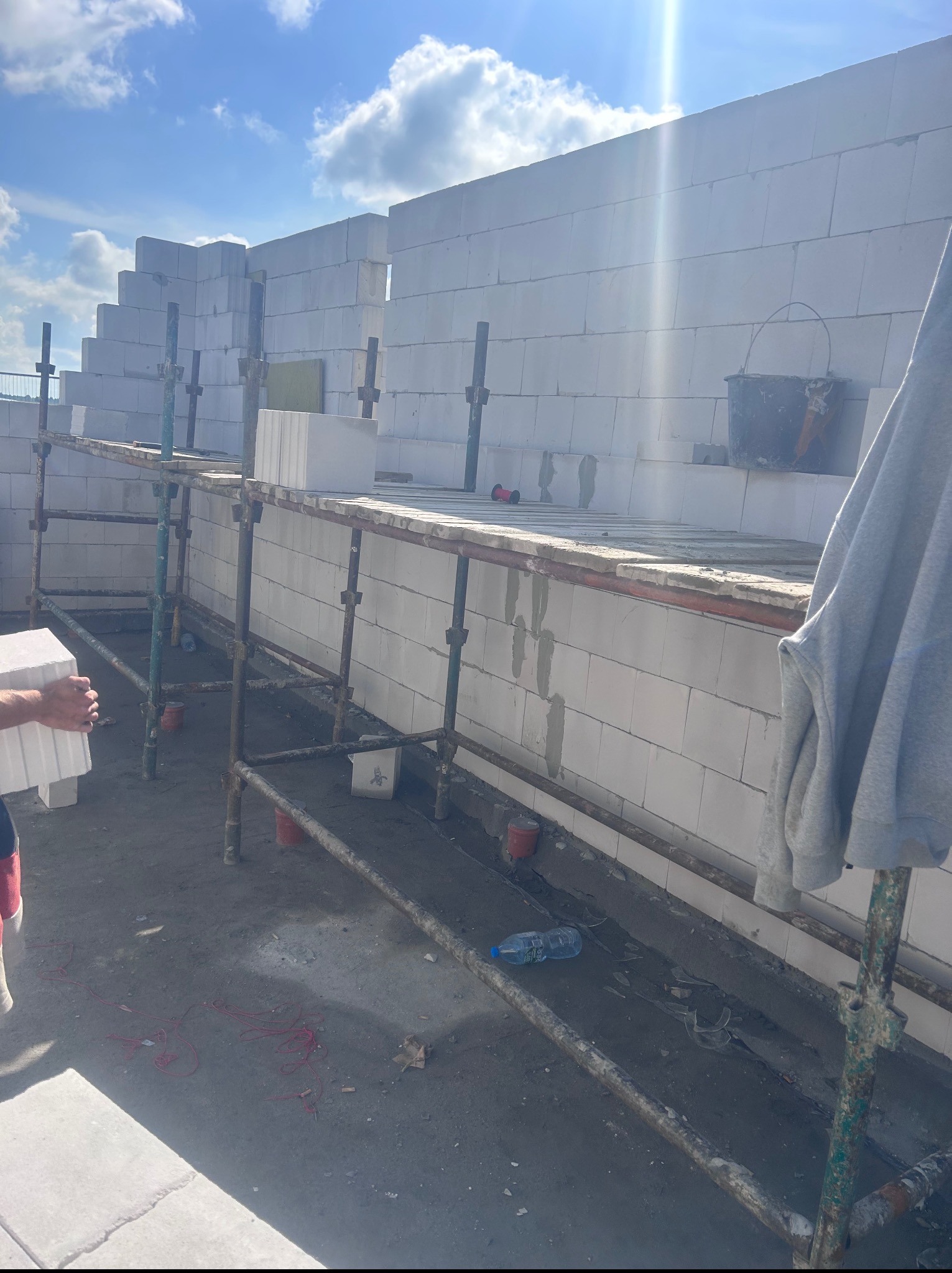 Wznoszenie ściany z białych bloczków na rusztowaniu. Widoczny fragment ręki z bloczkiem, wiadro i butelka. Słoneczny dzień, budowa w toku.