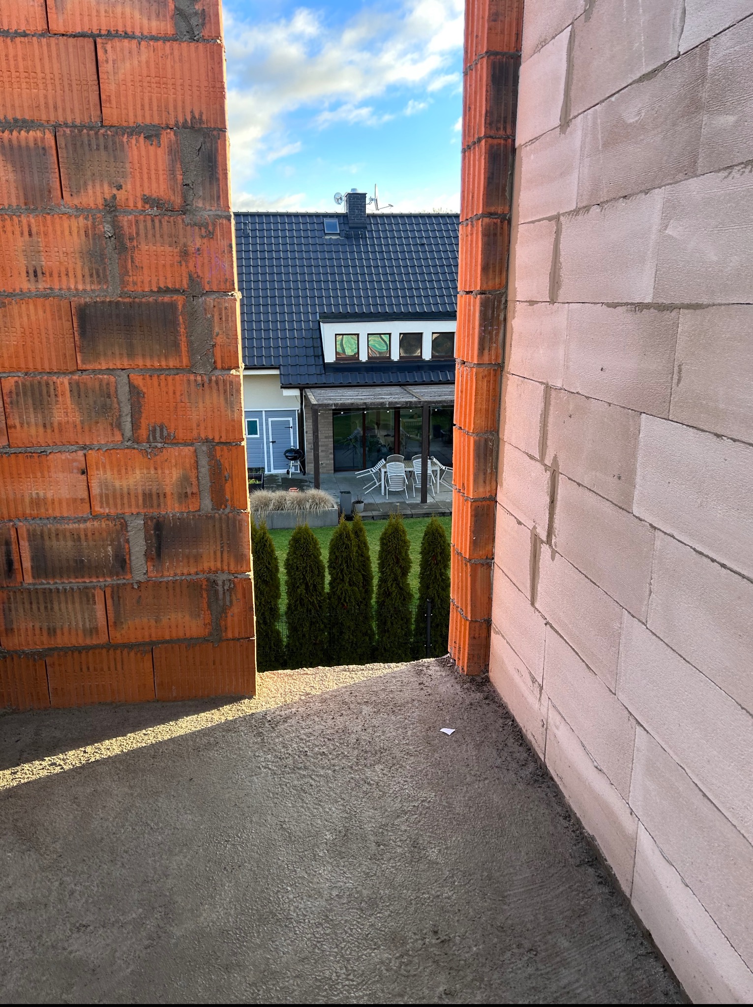 Wznoszenie ścian domu z pustaków ceramicznych i betonu komórkowego. Widoczny fragment ogrodu z tujami i budynek w tle. Wąskie ujęcie, prezentujące detale konstrukcji.