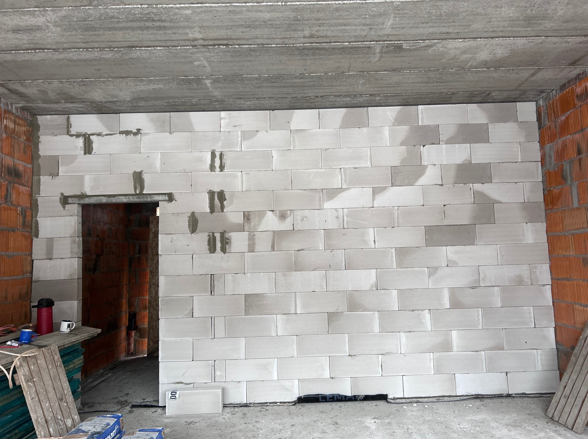 Ściana z białych bloczków, częściowo wymurowana, widoczne spoiny. W tle fragment ściany z cegły dziurawki i surowy betonowy strop. Na podłodze materiały budowlane.