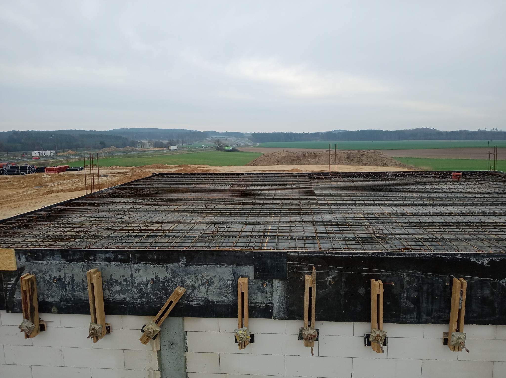 Zbrojenie płyty fundamentowej na budowie domu z bloczków, widok z góry na siatkę zbrojeniową, drewniane podpory i odległy krajobraz.