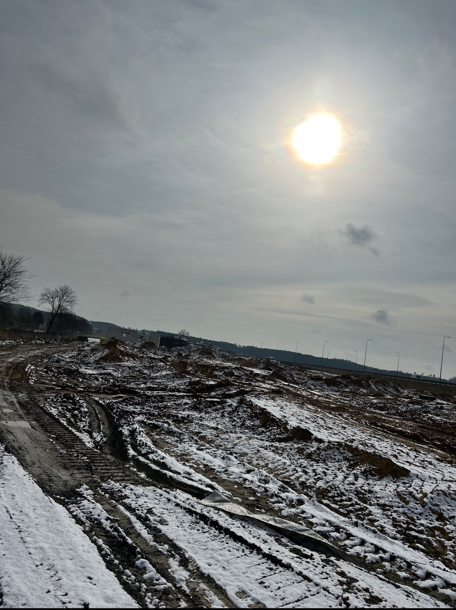 Zimowy krajobraz placu budowy z widocznymi śladami opon w błocie i śniegu, w tle wznoszona konstrukcja budynku oraz linia oświetlenia ulicznego.
