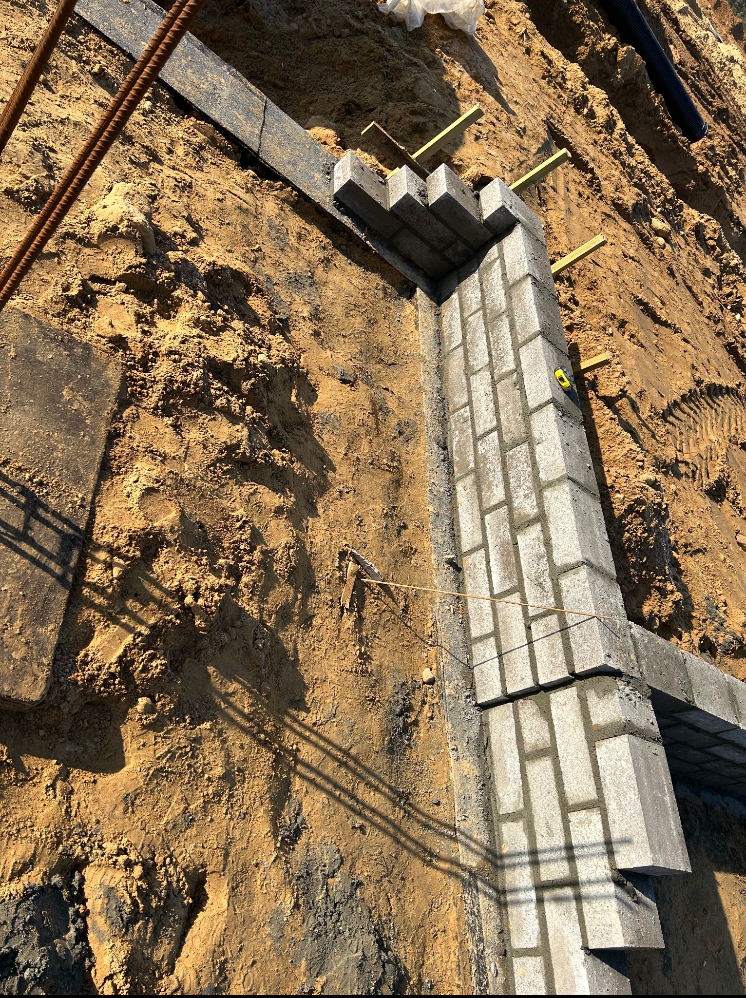 Wykop z częściowo wymurowanym fundamentem z szarych bloczków betonowych, zabezpieczonym deskami i poziomnicą. Widoczne pręty zbrojeniowe i ślady opon w ziemi.