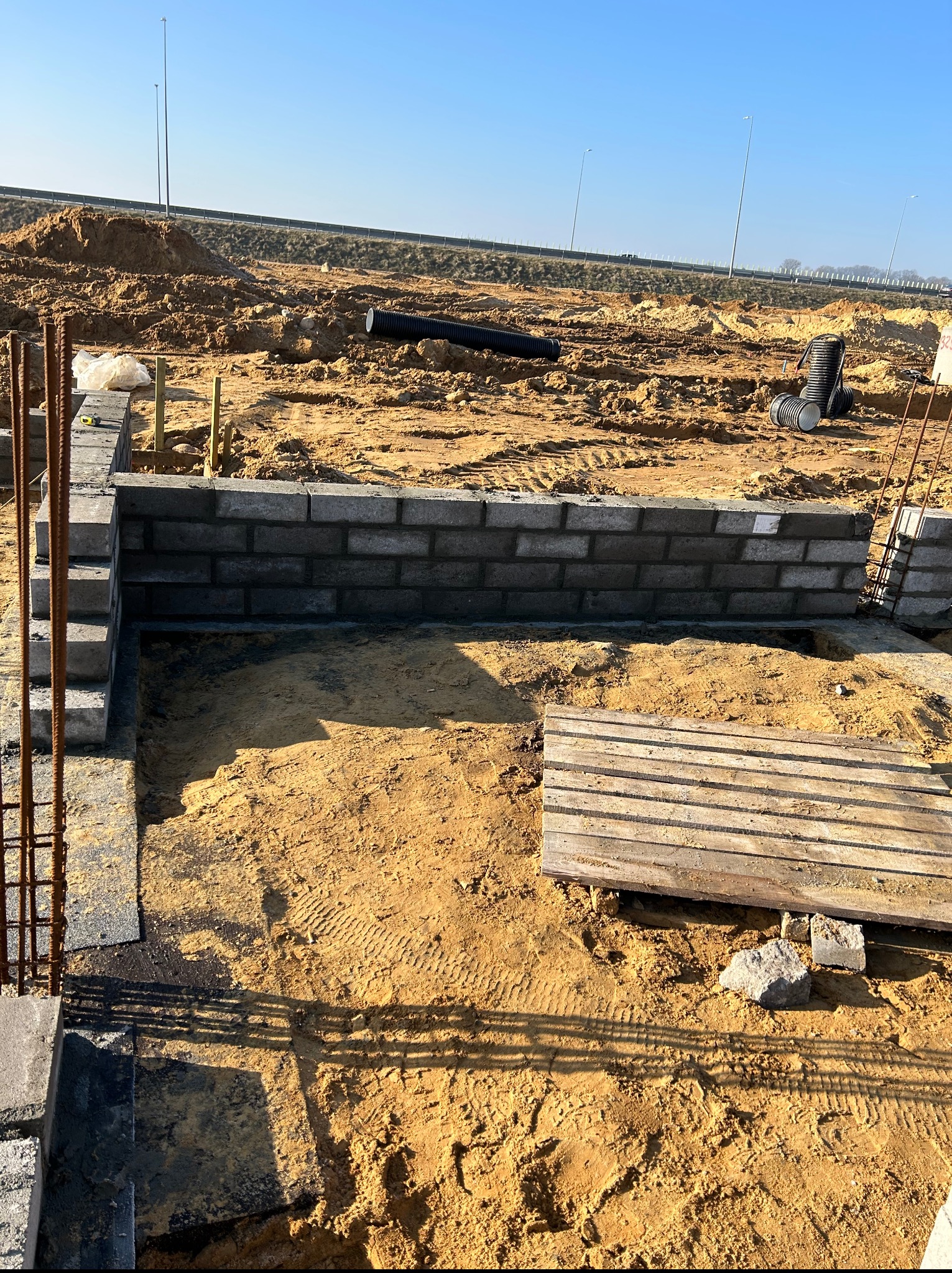Wykop pod fundamenty z częściowo wymurowanymi ścianami z bloczków betonowych. Widoczne zbrojenia i ślady opon w piasku. W tle krajobraz budowy i błękitne niebo.