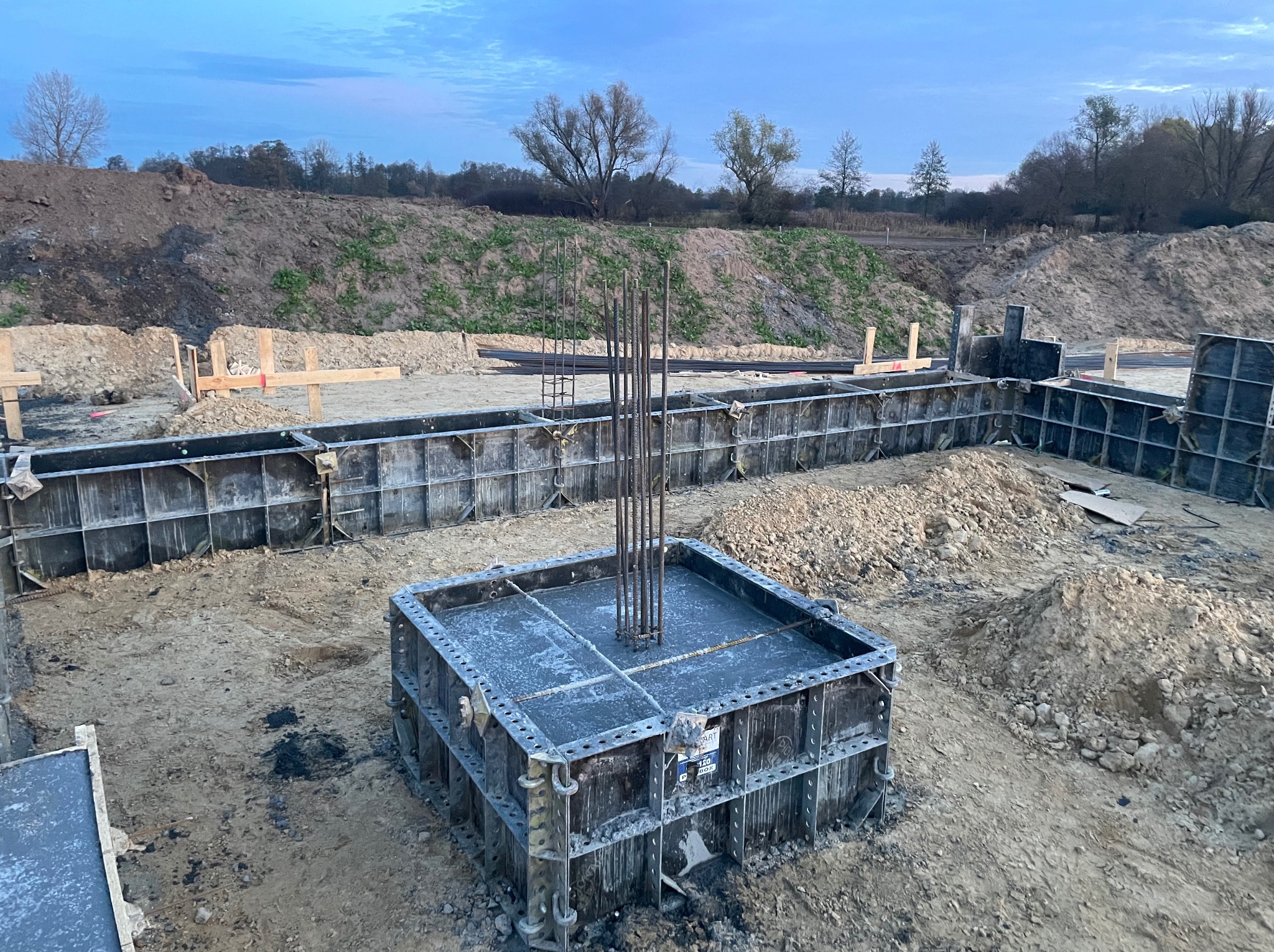 Zbrojenie fundamentu w szalunku systemowym, wystające pręty pionowe, teren budowy z ziemią i formami szalunkowymi, pochmurne niebo w tle.