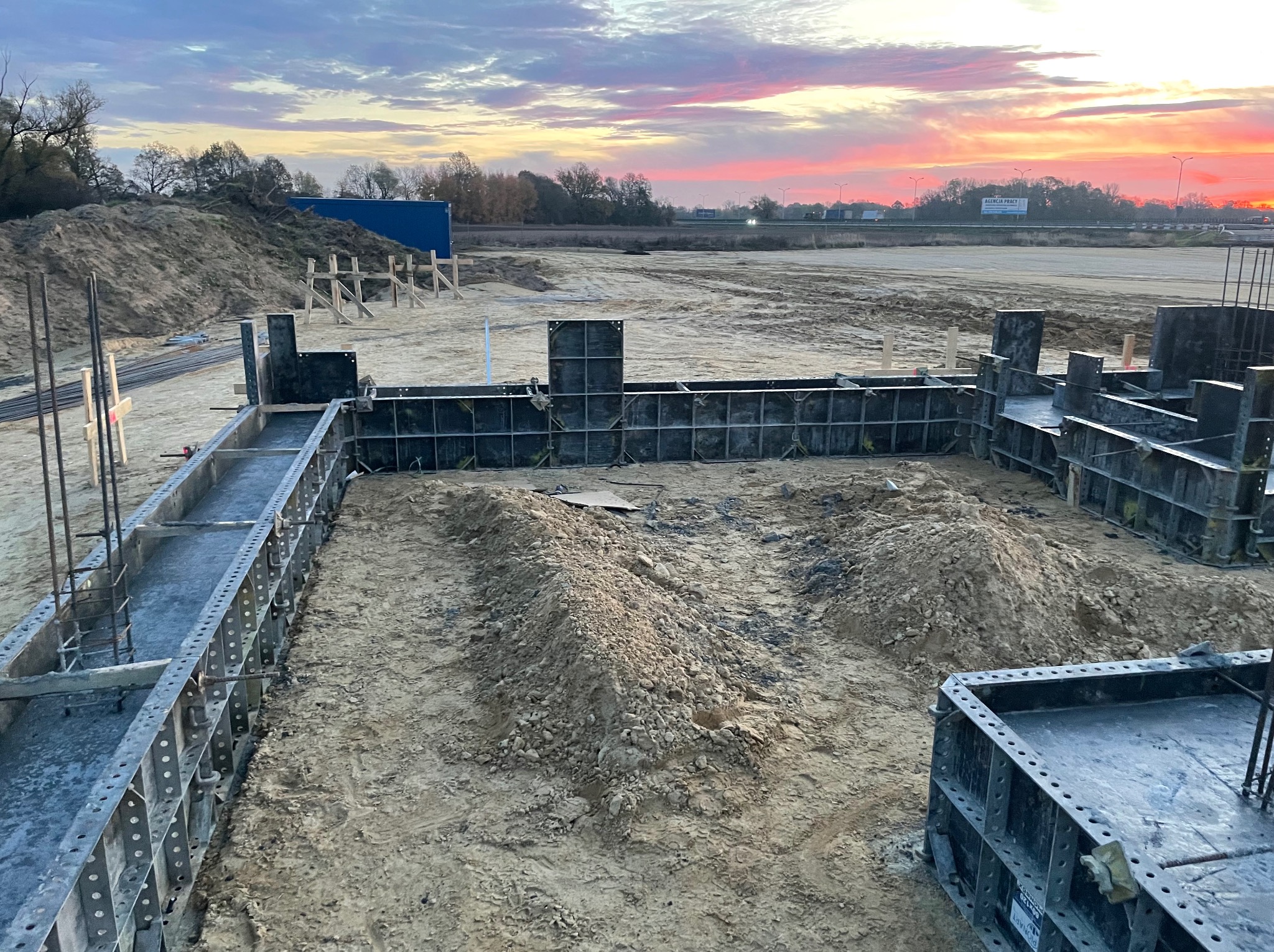 Zalewanie fundamentów w szalunkach na placu budowy, widok z góry, w tle zachodzące słońce i błękitny kontener. Etap wylewania betonu.