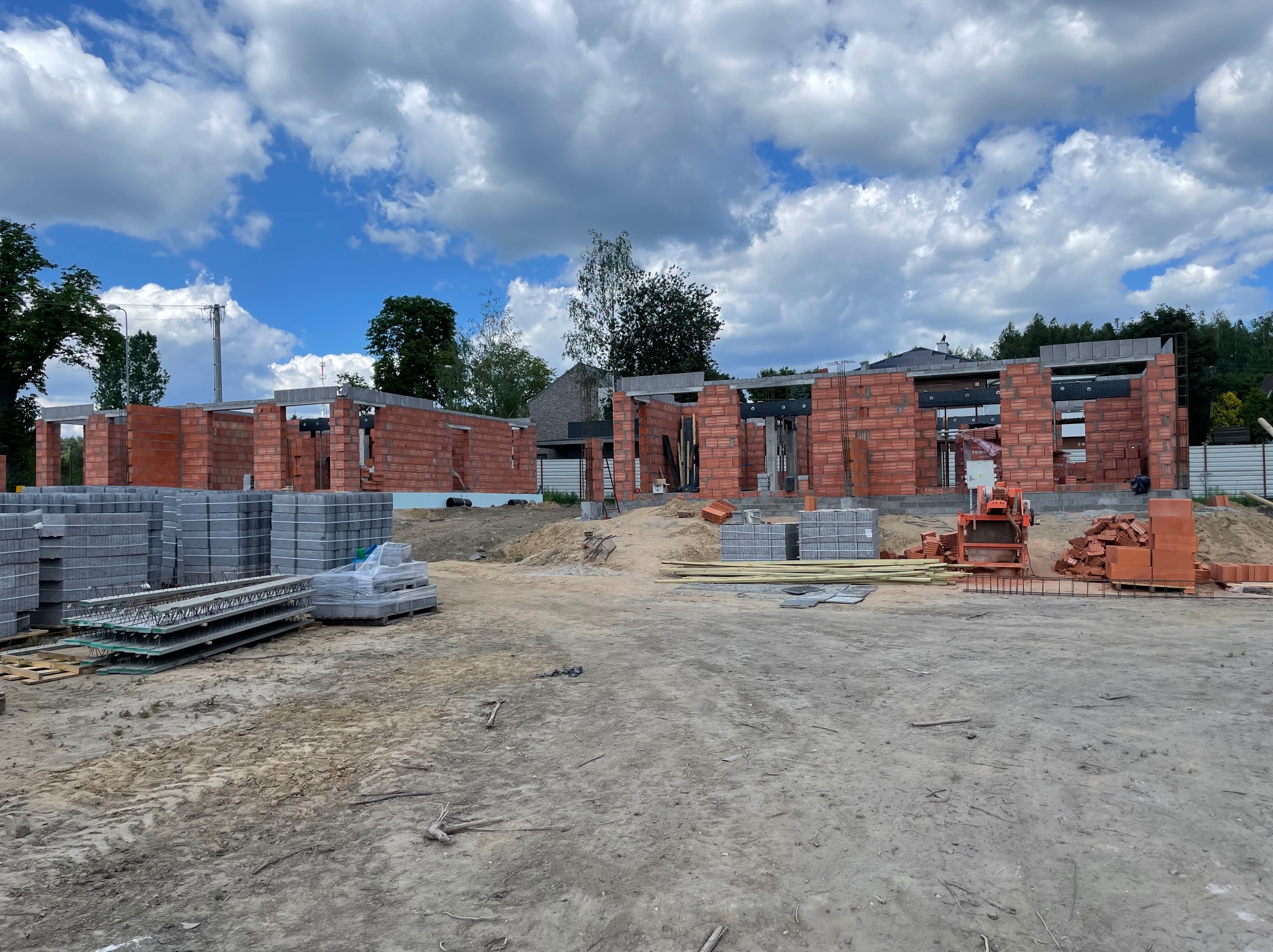 Wznoszenie murów z czerwonej cegły na budowie domu jednorodzinnego. Widoczne bloczki betonowe i elementy konstrukcyjne na tle błękitnego nieba z chmurami.