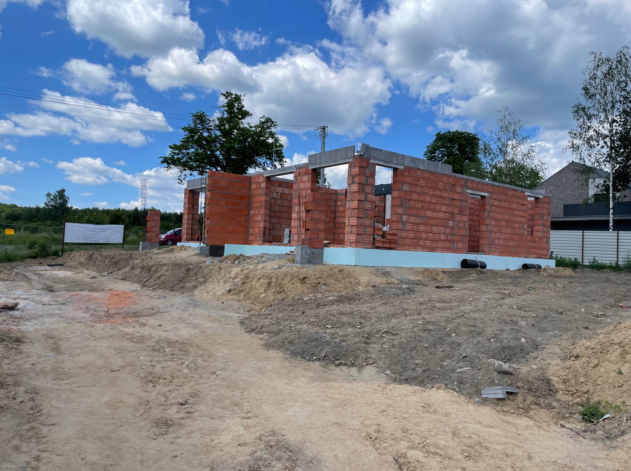 Budowa domu z czerwonej cegły na fundamencie z izolacją, widok z zewnątrz, w tle błękitne niebo z chmurami i zieleń drzew.