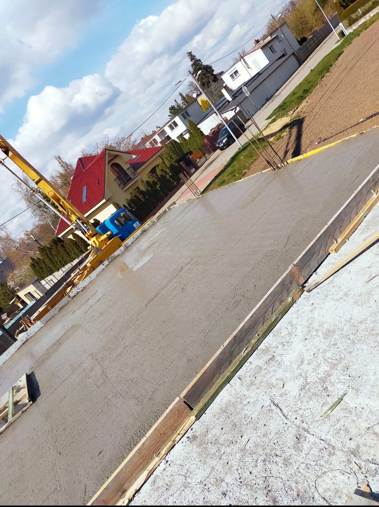 Betonowanie fundamentu domu jednorodzinnego z użyciem pompy do betonu. Widoczne szalunki i zbrojenie. W tle zabudowa mieszkalna i błękitne niebo.