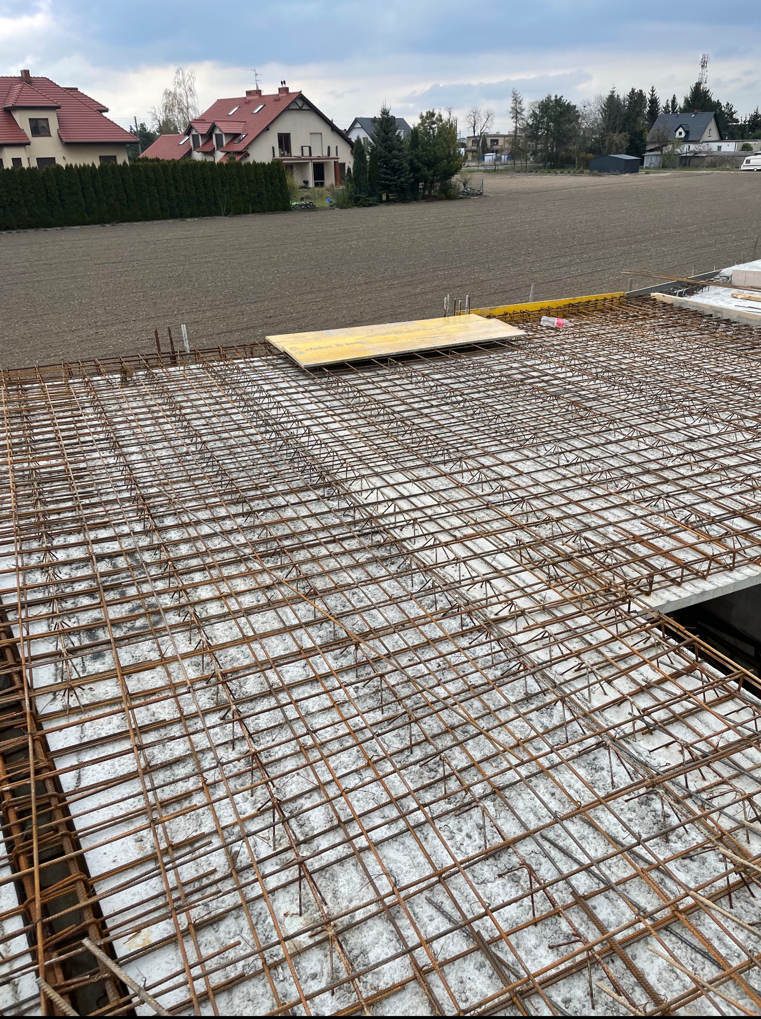 Zbrojenie płyty fundamentowej na styropianie, widok z góry. Na drugim planie zabudowa jednorodzinna i pole uprawne. Szary dzień.