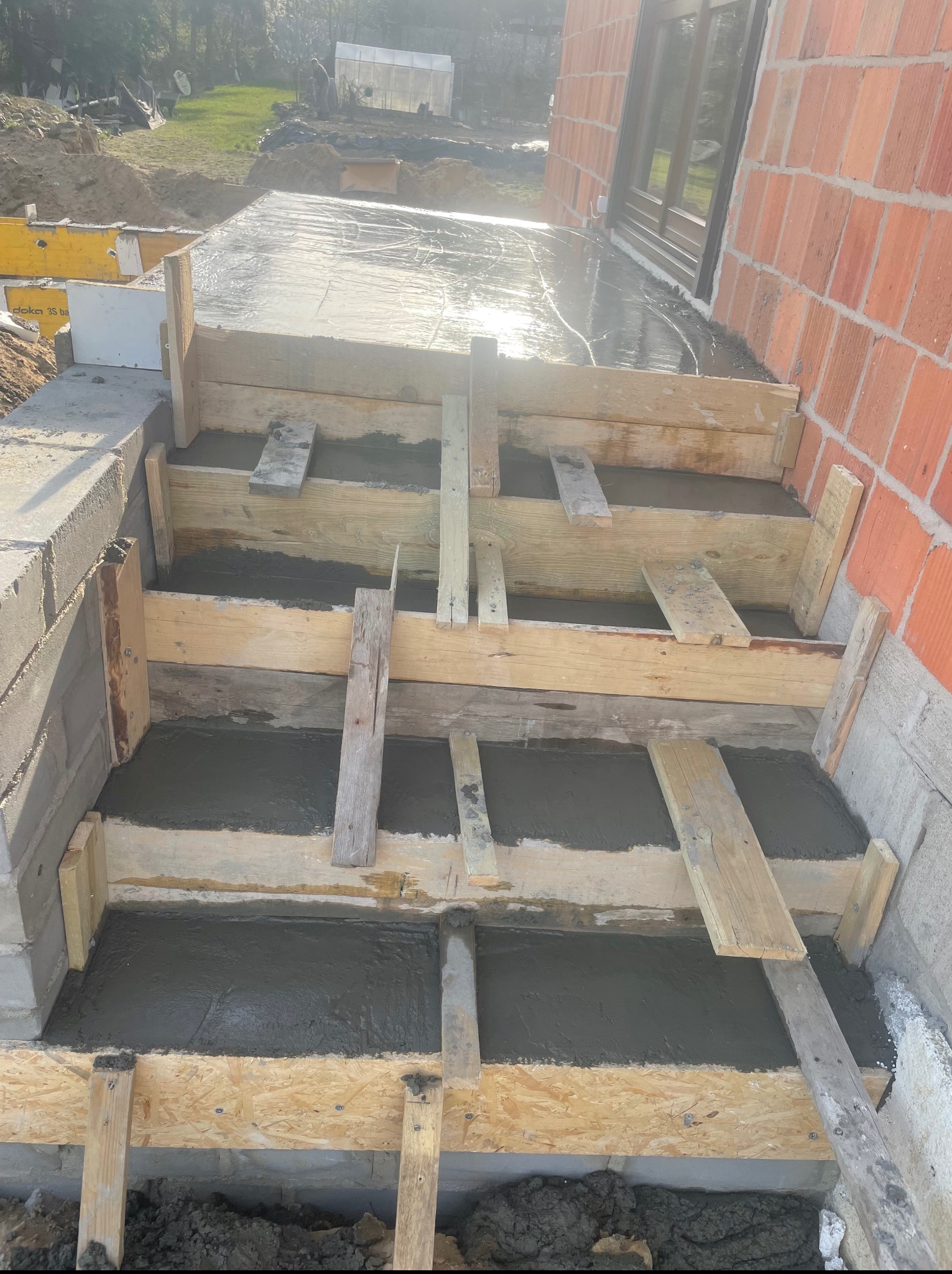 Wylewka betonowa schodów wejściowych do domu z cegły, szalunek z desek i płyt OSB, widok z góry na plac budowy w słoneczny dzień.