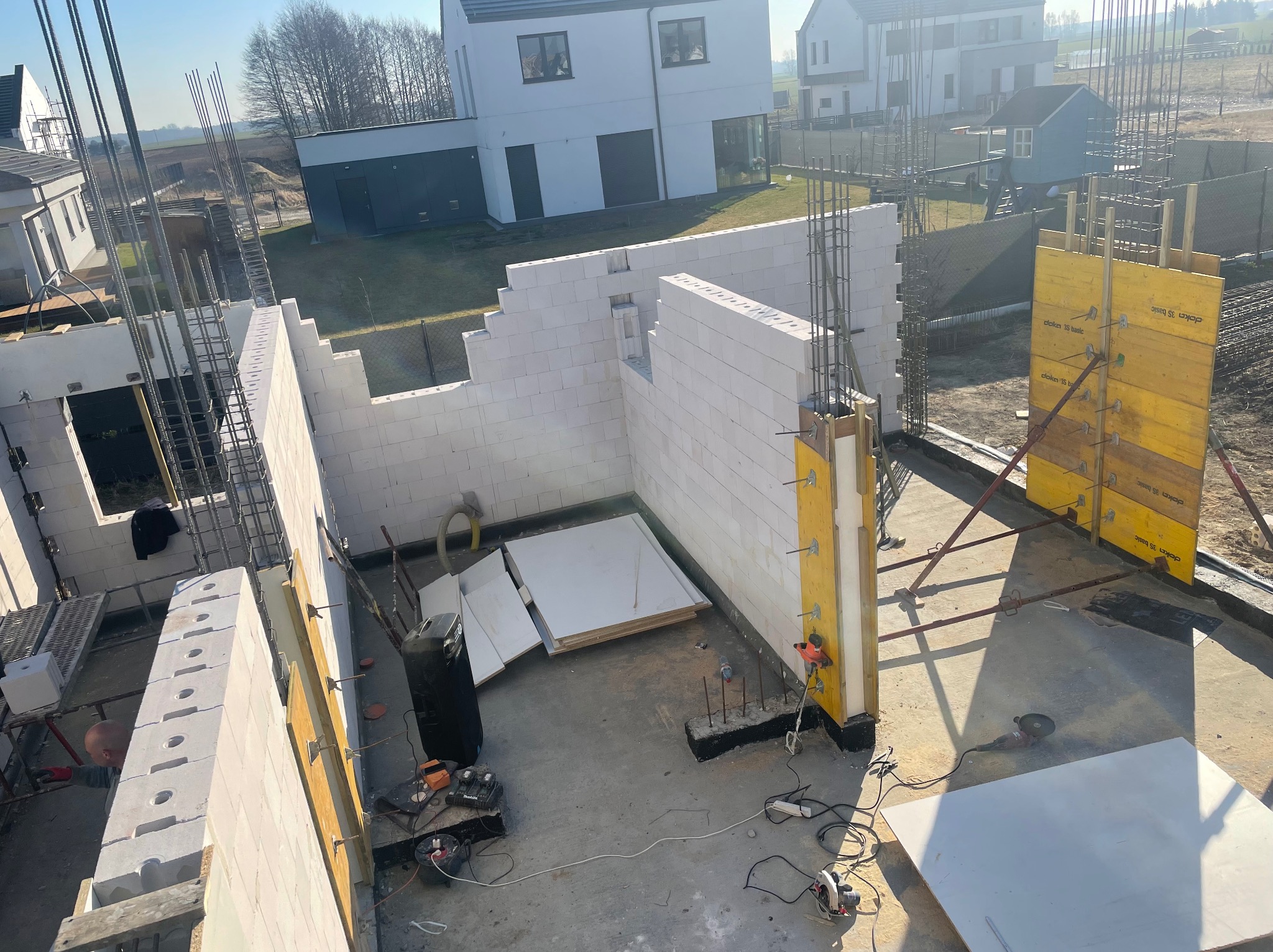 Budowa domu z betonu komórkowego: wzniesione ściany, widoczne zbrojenie i szalunki. Na podłodze narzędzia budowlane, w tle domy jednorodzinne.