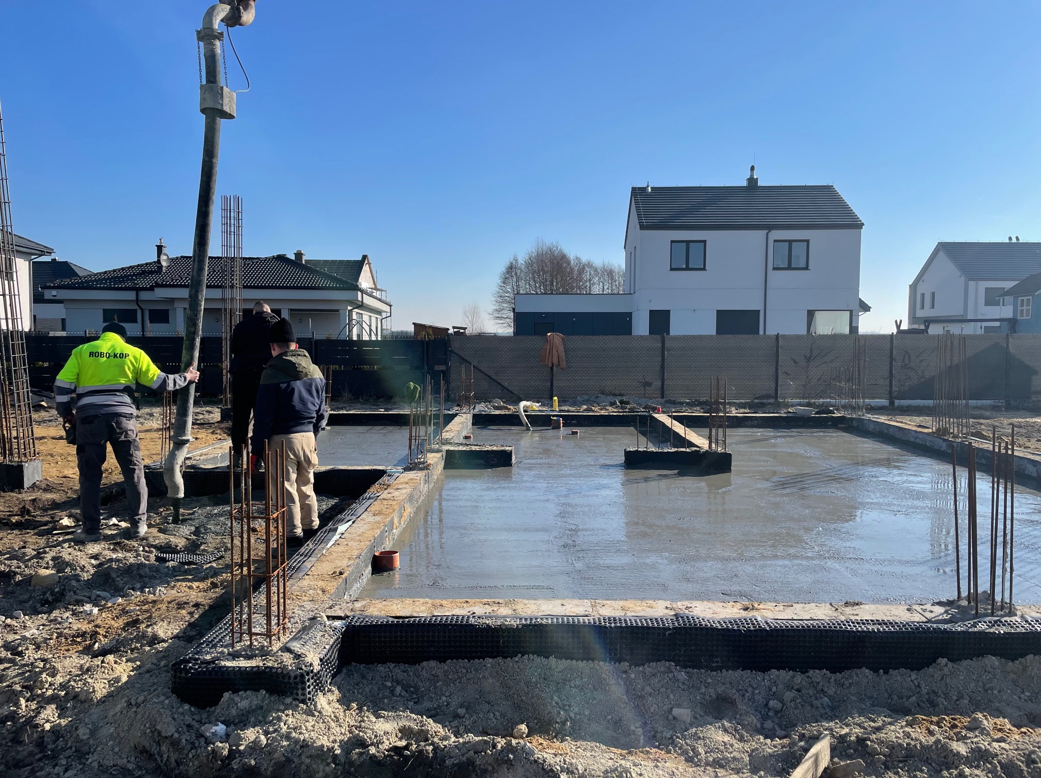 Wylewka fundamentów domu jednorodzinnego w słoneczny dzień. Widoczne zbrojenia, pracownicy i pompa do betonu. W tle zabudowa jednorodzinna.