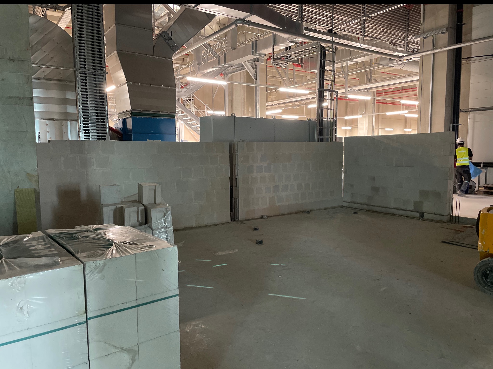 Wnętrze hali w trakcie budowy: częściowo wymurowane ściany z bloczków, palety z materiałem budowlanym, instalacje pod sufitem, pracownik w oddali.