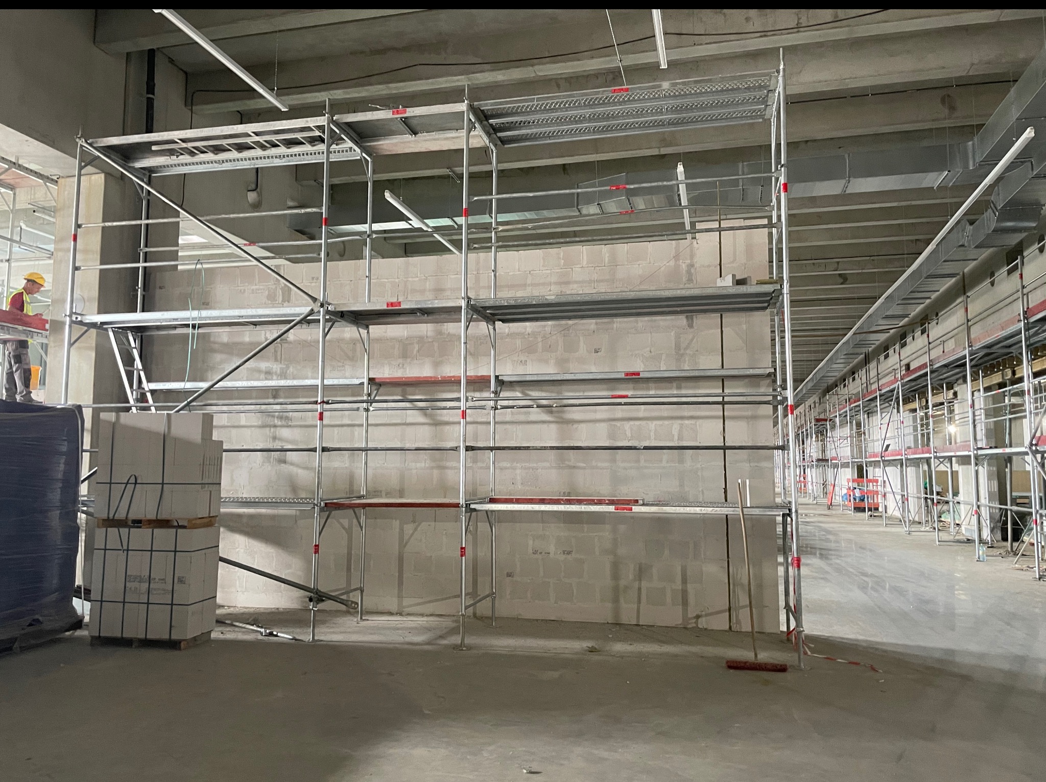 Wnętrze w trakcie prac murarskich: ściana z bloczków, rusztowanie, paleta z materiałem. Pracownik w oddali. Widok na plac budowy w surowym stanie.