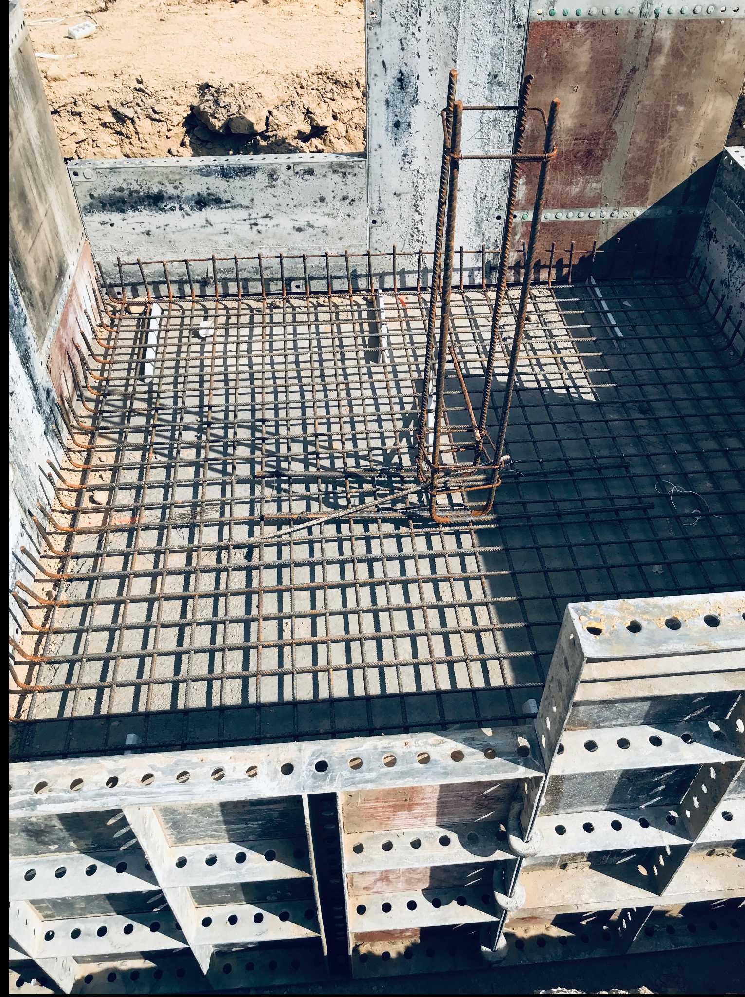 Zbrojenie fundamentu w szalunku stalowym, widok z góry. Stalowe pręty tworzą siatkę, a pionowe elementy zbrojenia wystają ponad nią. Część szalunku widoczna w dolnej części kadru.