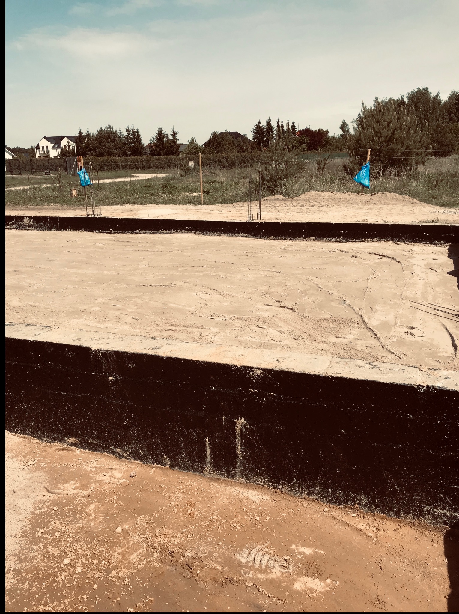 Zabezpieczone fundamenty domu jednorodzinnego z wystającymi prętami zbrojeniowymi, widok z góry, w tle zabudowa jednorodzinna i zieleń.