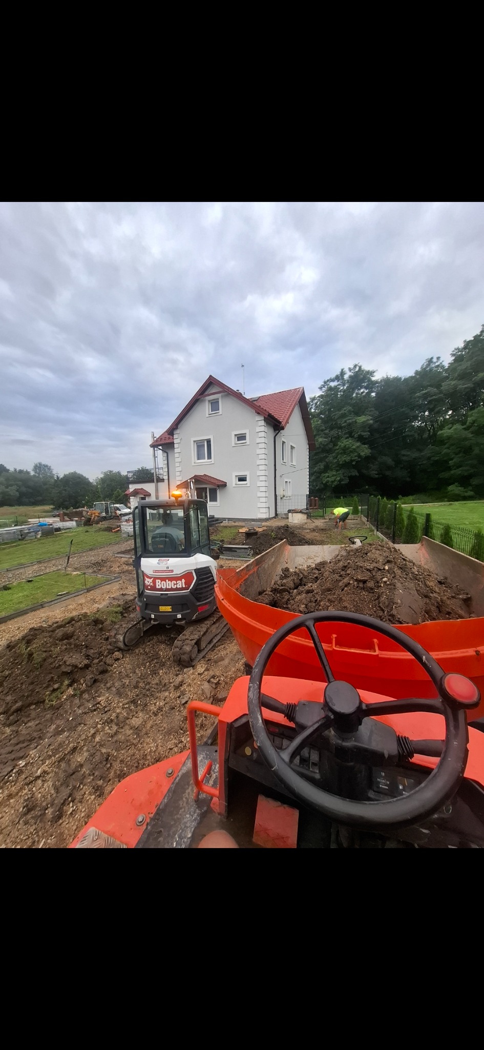 Bobcat i wywrotka z ziemią na posesji przed domem. Przygotowanie terenu pod ogród. Prace ziemne w toku. Widok z perspektywy operatora wywrotki.