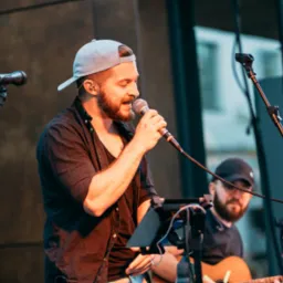 Wokalista z brodą śpiewa do mikrofonu, w tle gitarzysta. Scena z nagłośnieniem i oświetleniem, dynamiczne ujęcie z koncertu na żywo.