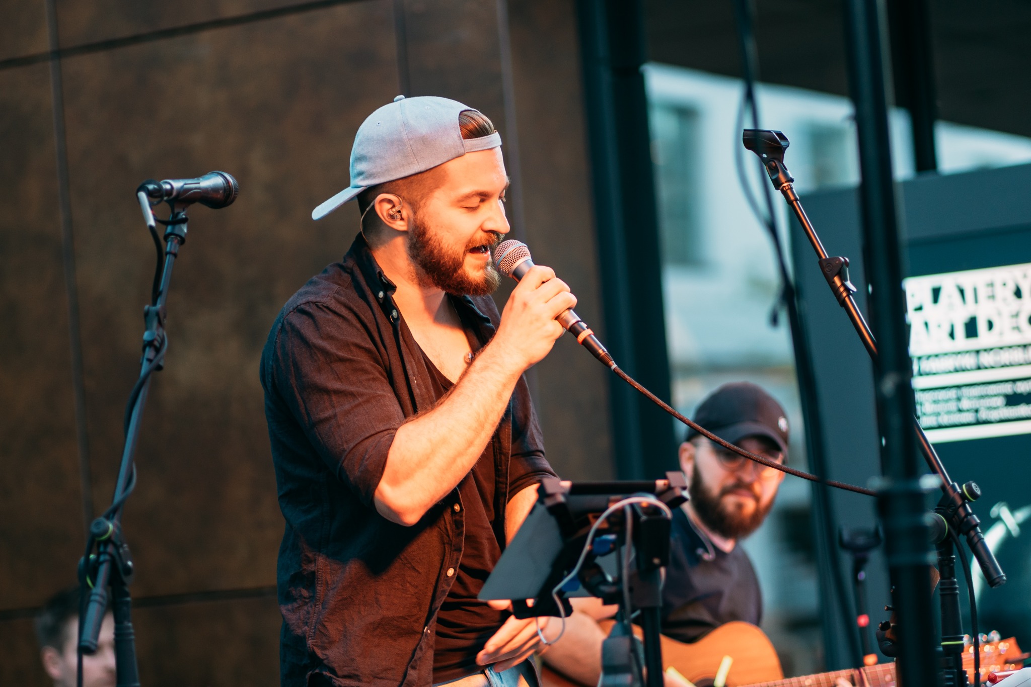 Wokalista z brodą śpiewa do mikrofonu, w tle gitarzysta. Scena z nagłośnieniem i oświetleniem, dynamiczne ujęcie z koncertu na żywo.