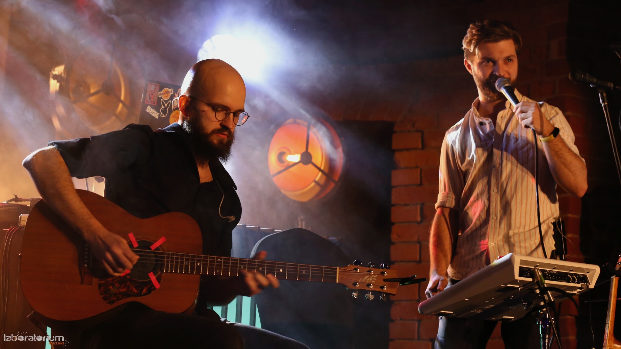 Duet muzyczny na scenie: gitarzysta z gitarą oklejoną taśmą i wokalista z mikrofonem przy klawiszach, ceglana ściana i reflektory tworzą klimat koncertu.