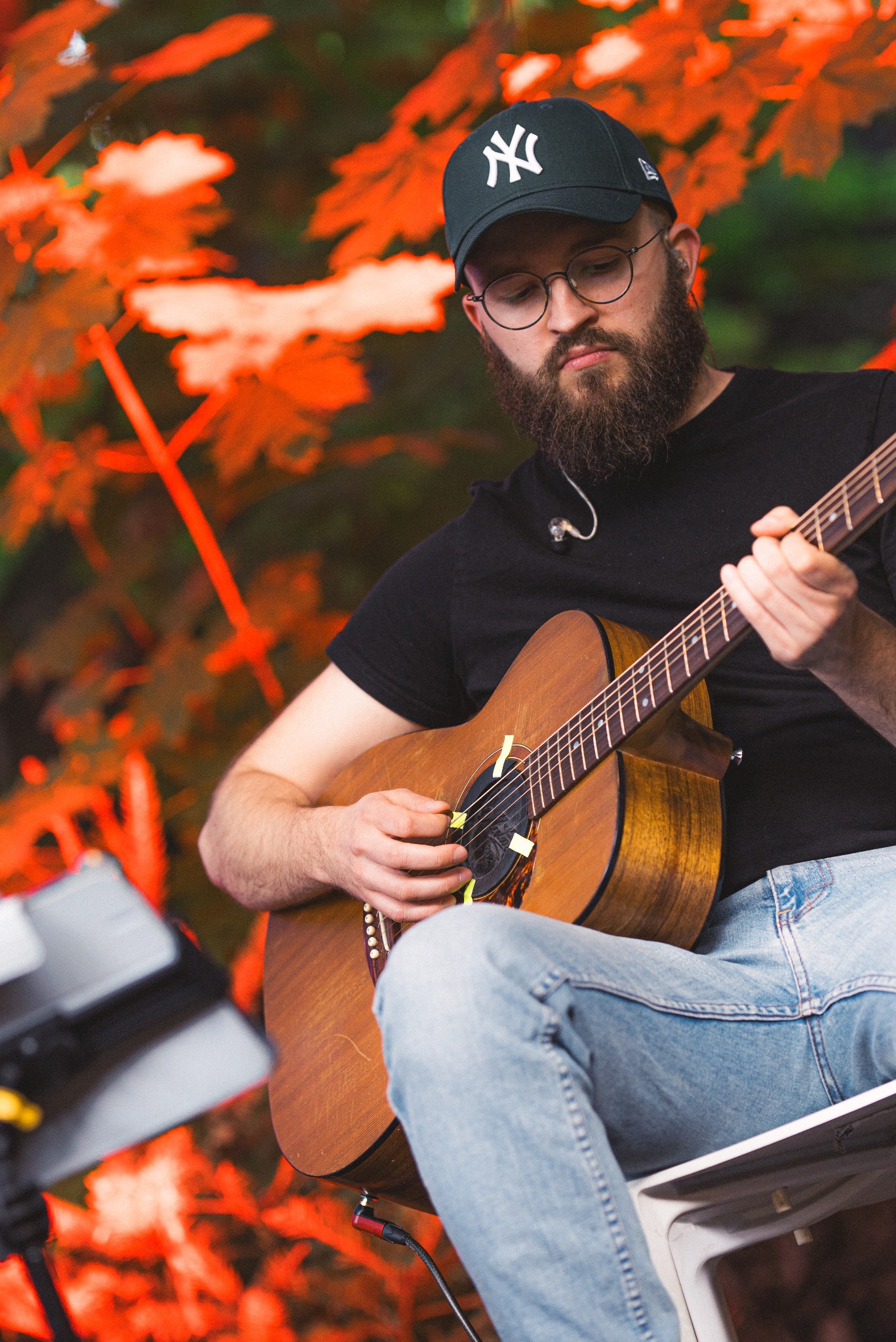 Gitarzysta z brodą w czarnej koszulce i czapce gra na akustycznej gitarze na tle pomarańczowych liści. Widoczne naklejki na gitarze i przewód.